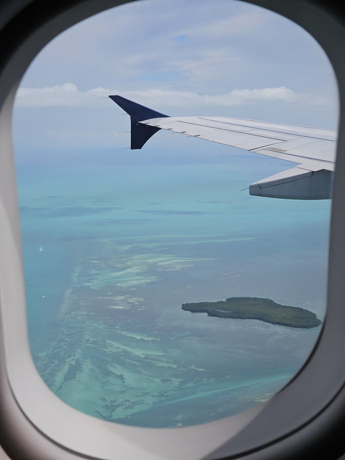 View of a tropical ocean and island from an airplane window, showcasing turquoise waters and clear skies.
