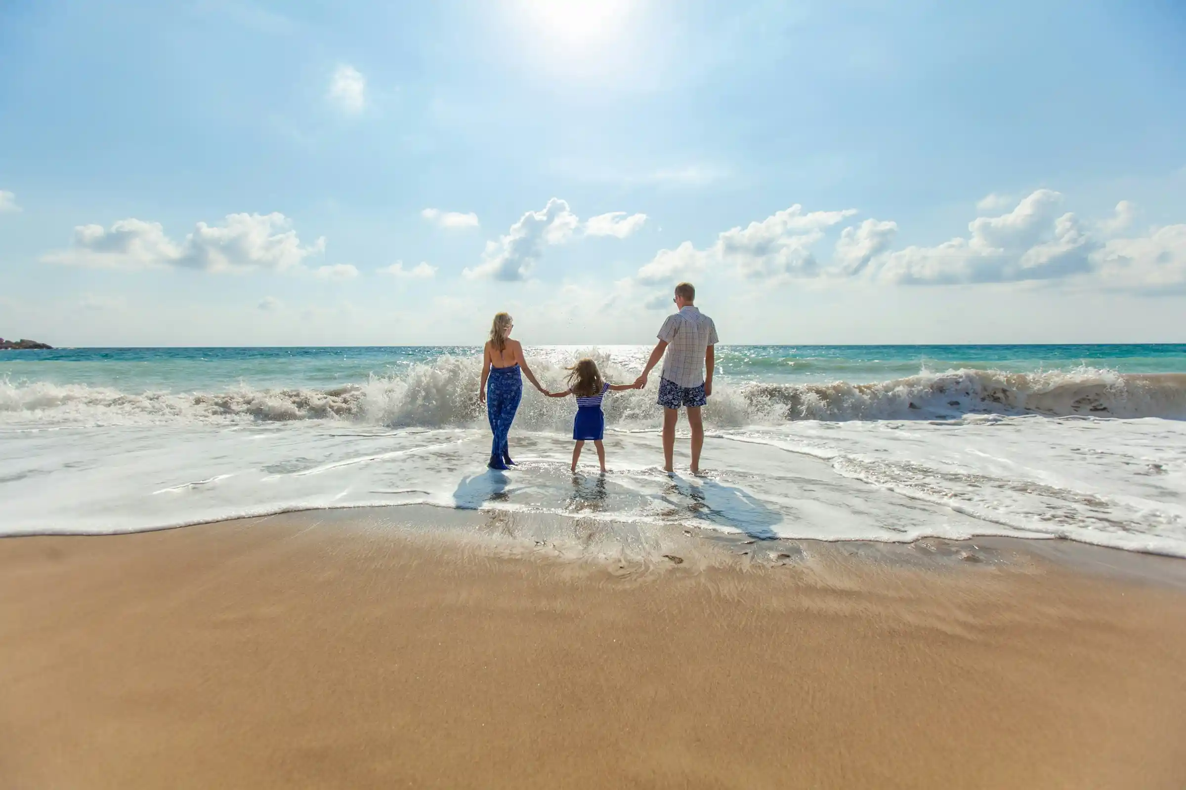 Parents and a child holding hands at the water's edge on a sunny beach