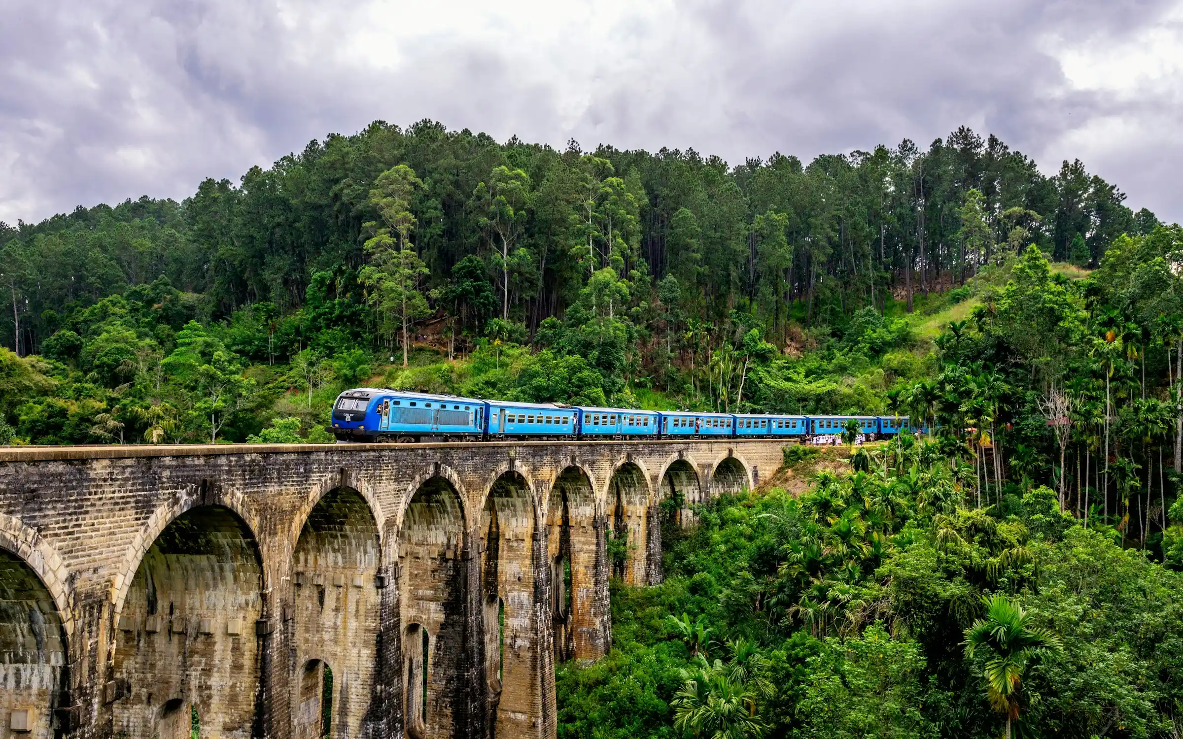 A blue train crossing the Nine Arch Bridge amid green hills in Ella, Sri Lanka