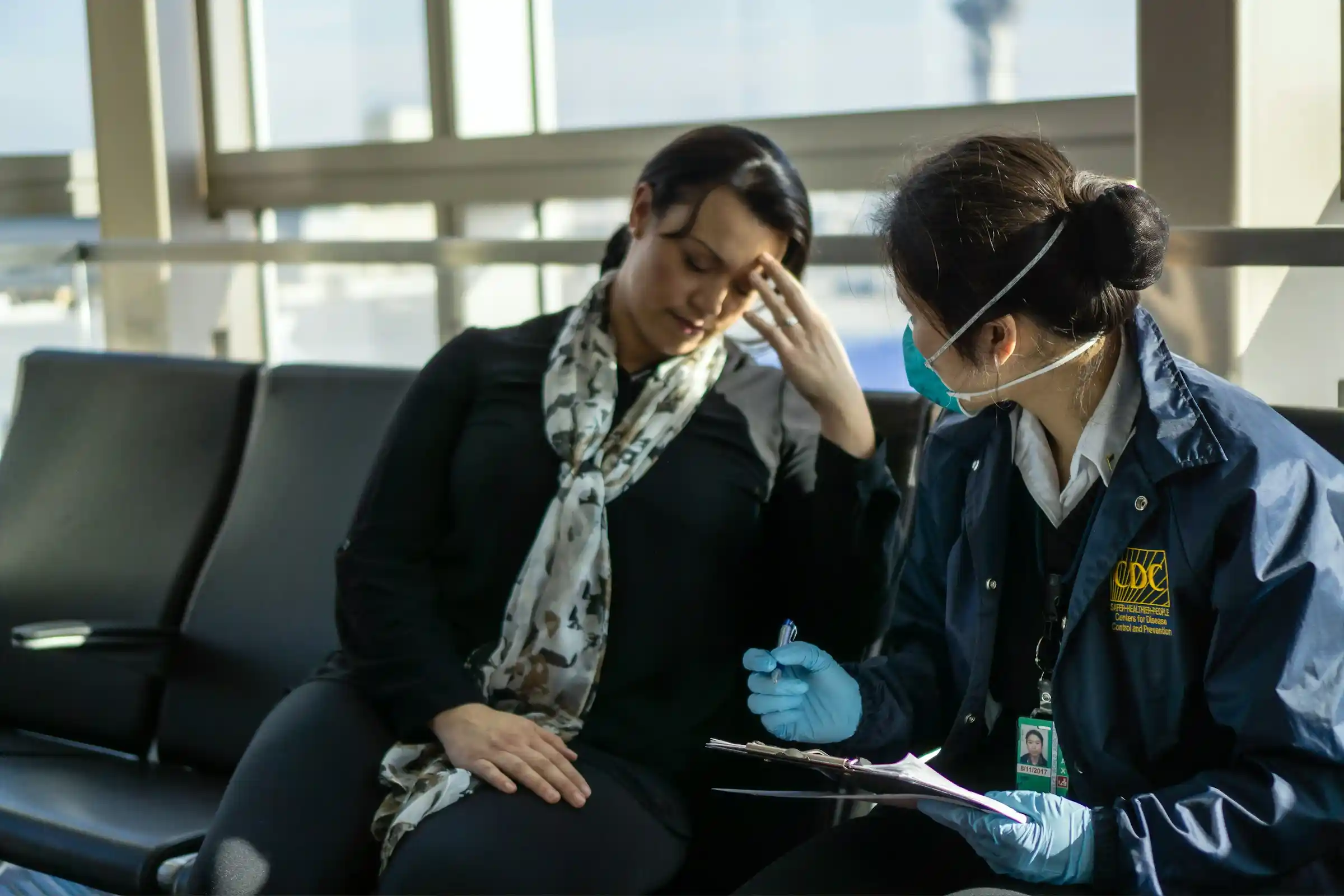 A masked health worker assisting an unwell traveller seated at an airport gate