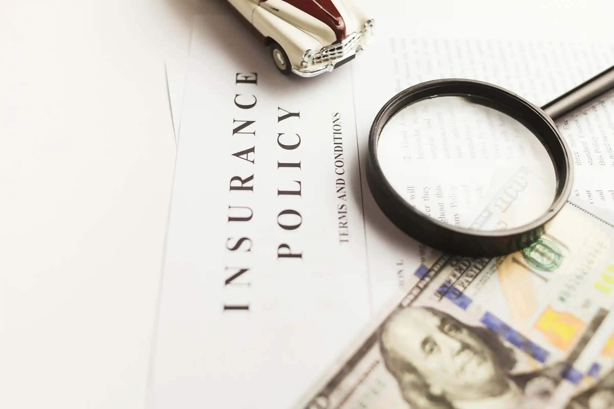 An insurance policy document under a magnifying glass beside cash and a toy car