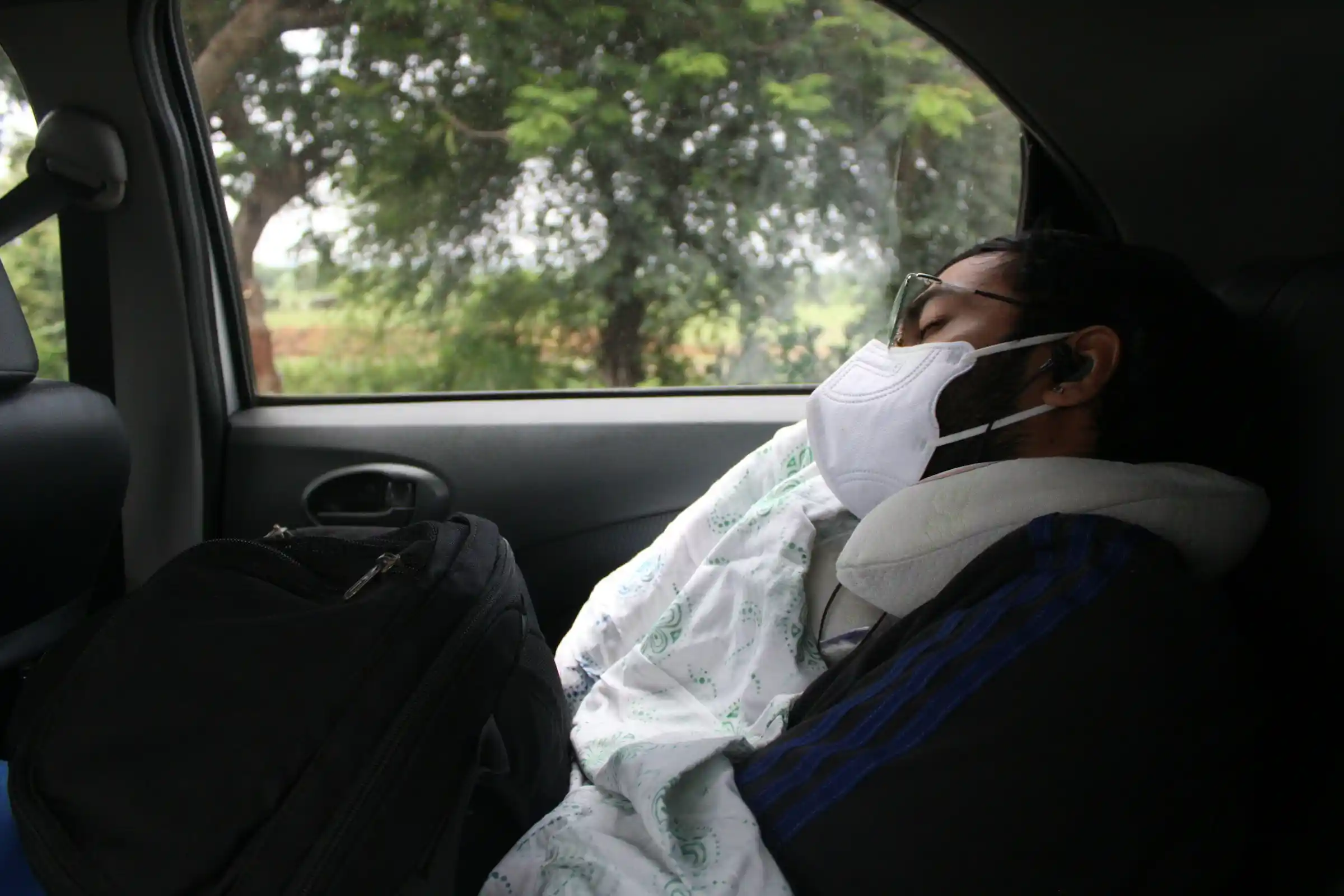 A tired traveller wearing a face mask and neck pillow resting in a car