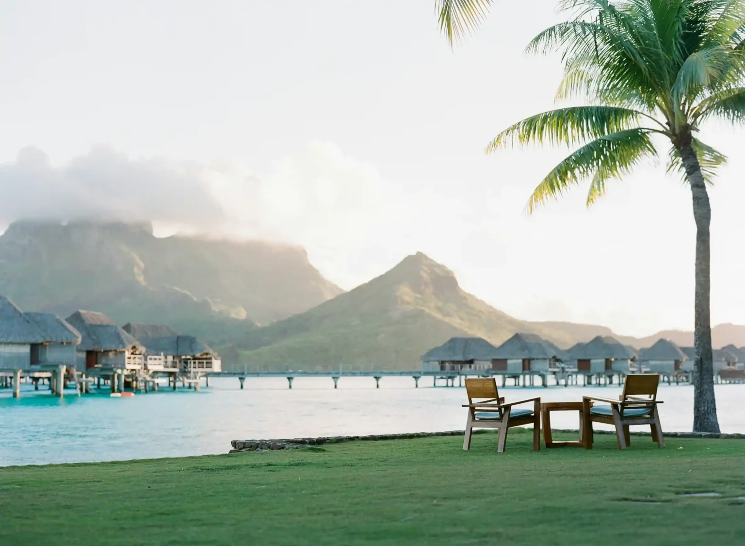 Overwater bungalows and a palm-lined resort lawn on the Bora Bora lagoon with the island's peaks behind