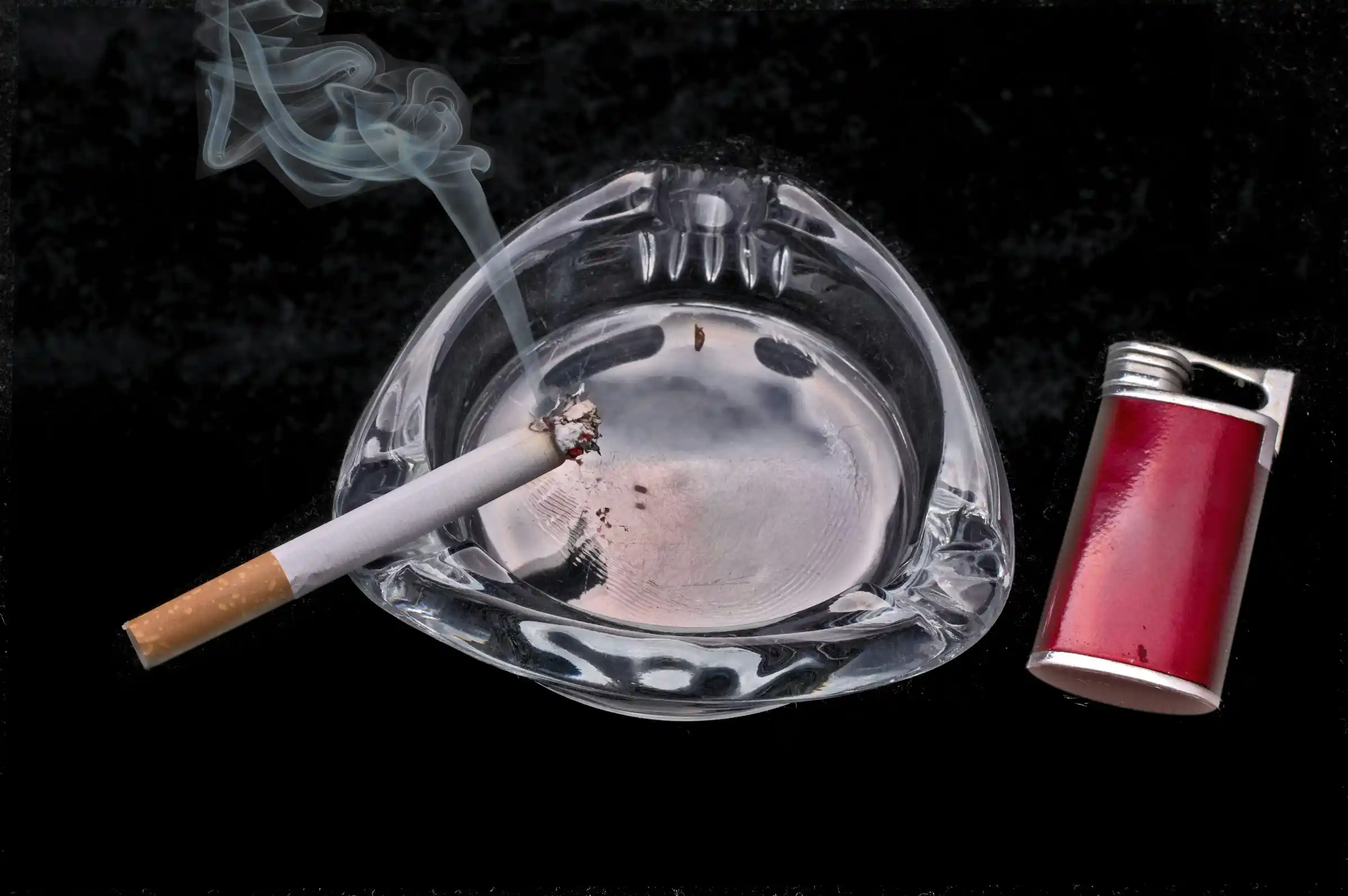 A lit cigarette resting in a glass ashtray beside a red disposable lighter