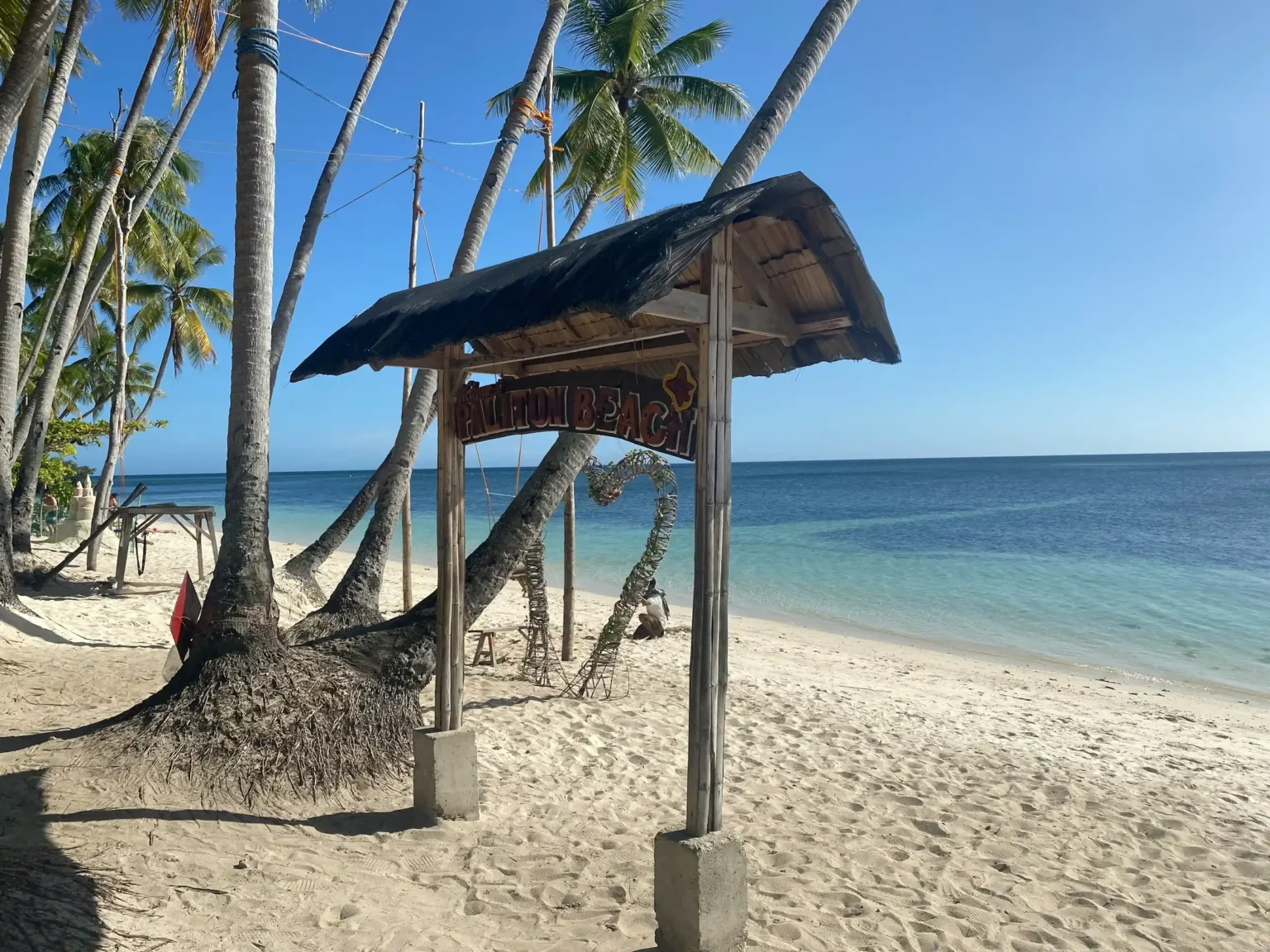 The bamboo Paliton Beach sign and leaning palm trees on the white sand of Paliton Beach, Siquijor
