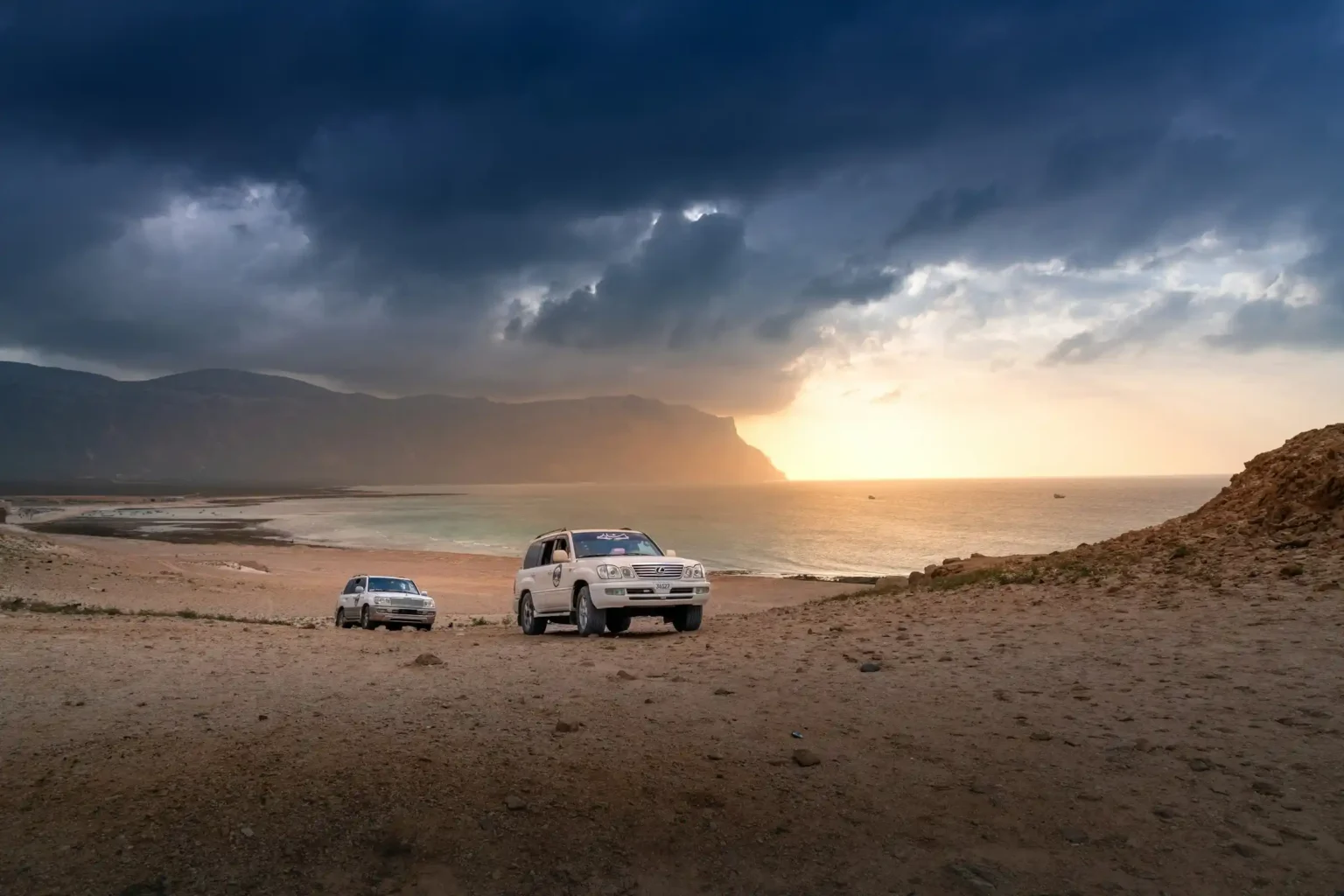 Two 4x4 vehicles on a rugged Socotra coastline at sunset with dramatic clouds