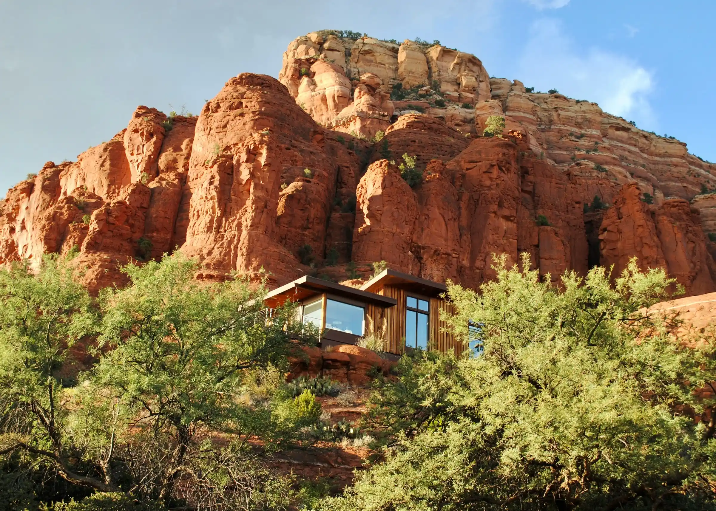 Modern glass cabin tucked into the base of a towering red rock butte among green trees in Sedona